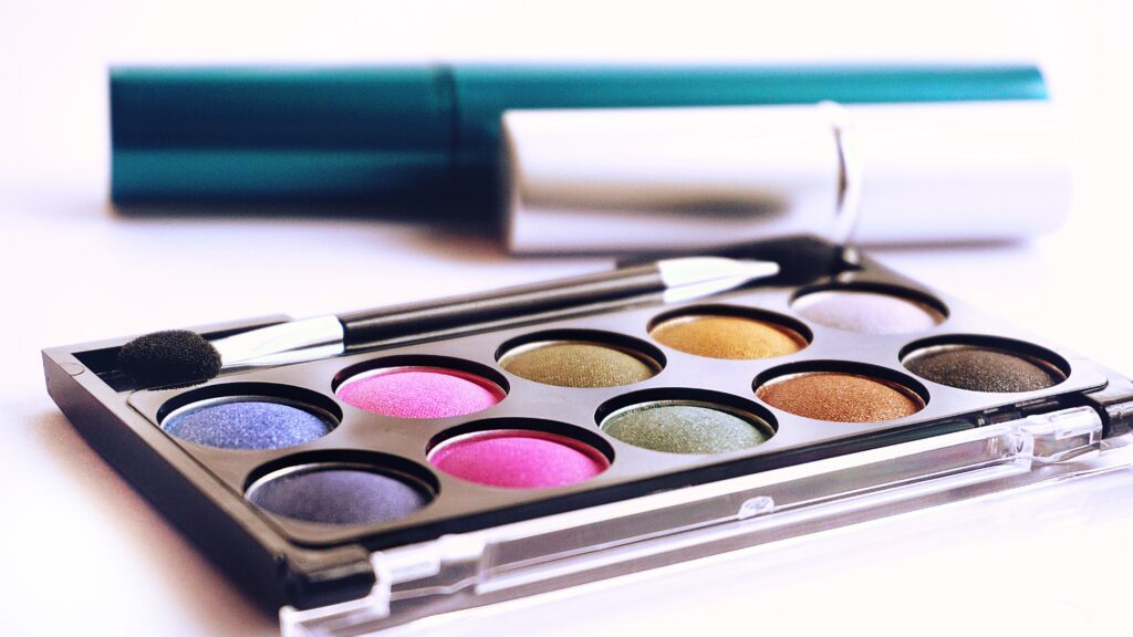 Close-up of a vibrant makeup palette featuring circular pans of shimmering purple, pink, gold, and green eyeshadows, with a double-ended applicator brush resting on top.