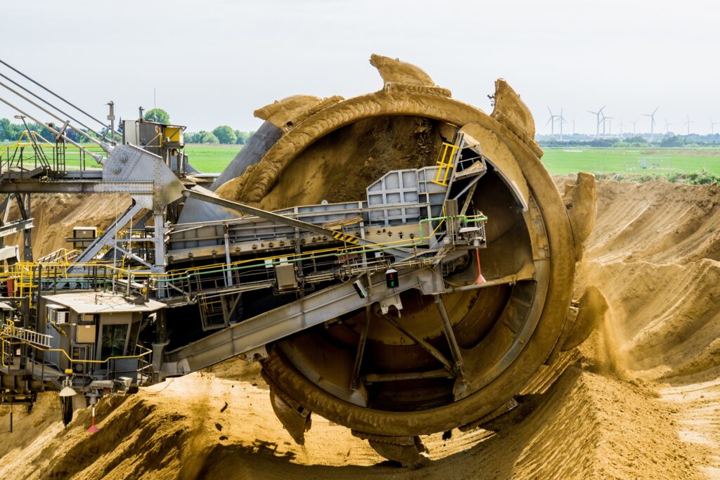 A massive industrial bucket-wheel excavator with a giant rotating wheel and multiple digging buckets, operating in an open-pit mine to move large quantities of earth and soil.