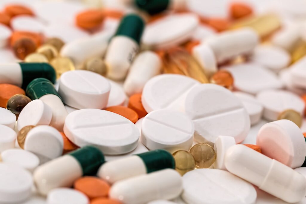 A close-up, top-down view of a variety of pharmaceutical pills and capsules, including round white tablets, orange-colored tablets, green and white capsules, and small translucent yellow gel caps.
