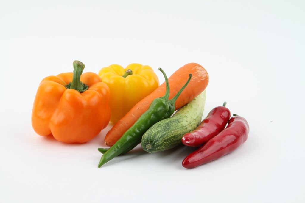 A vibrant arrangement of fresh vegetables on a plain white background, including an orange bell pepper, a yellow bell pepper, a whole carrot, a green chili, a cucumber, and two red chilies.